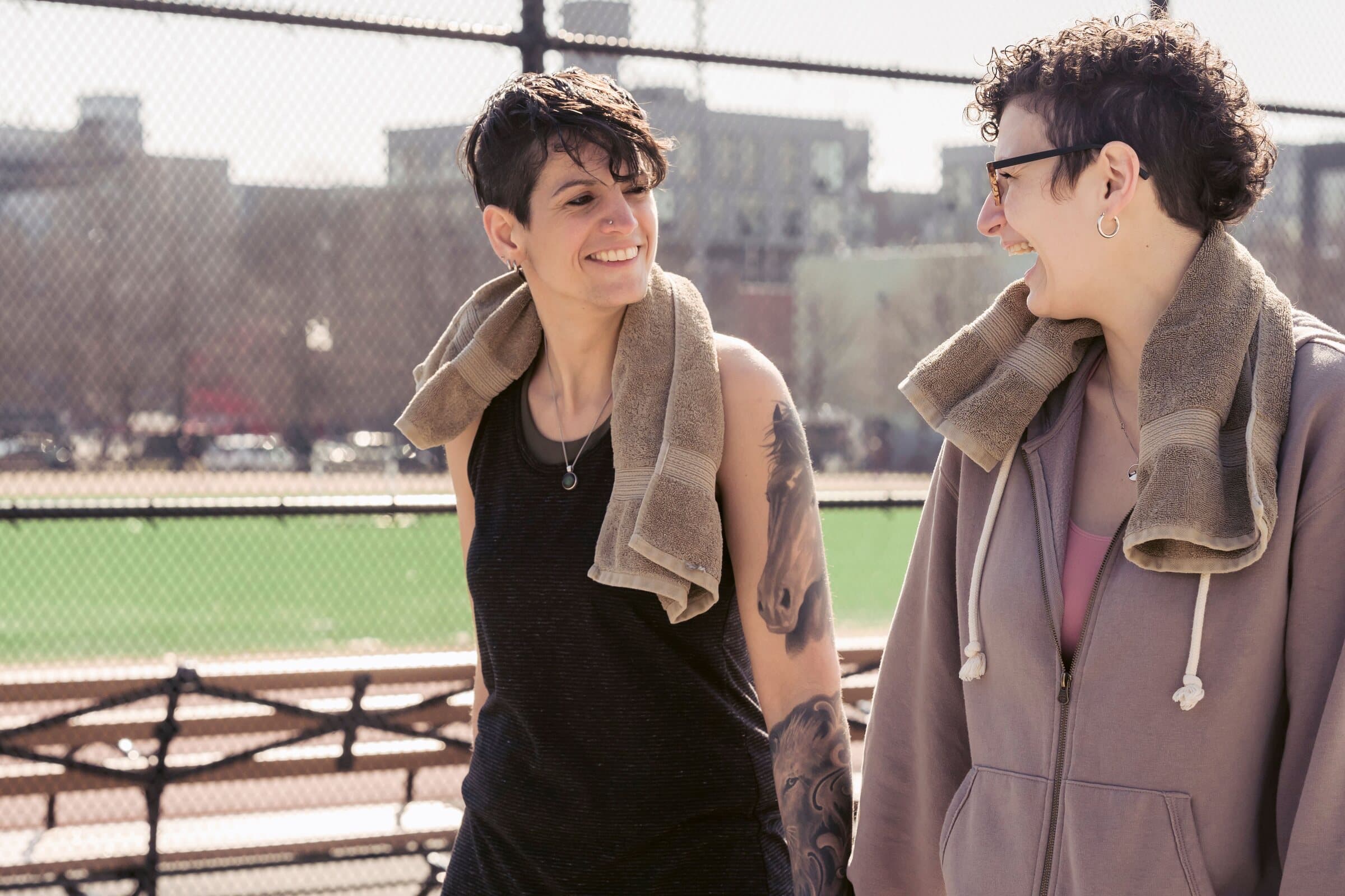 Two friends laughing together after exercise, towels around their necks, beside a city sports field